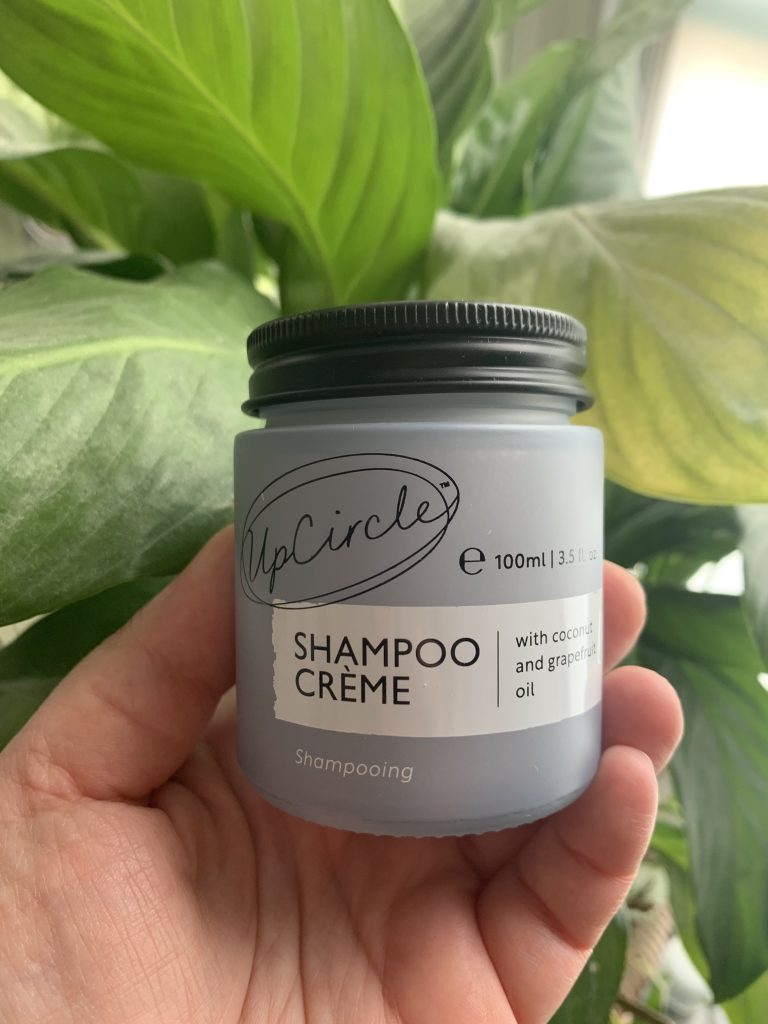 Shampoo creme by upCircle in a frosted pale bluee glass jar with a black metal lid. Set against a green leaf backdrop