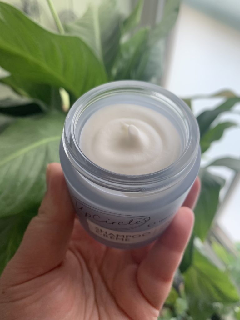 White, whipped shampoo creme pictured in a pale blue glass jar with a green leafy background