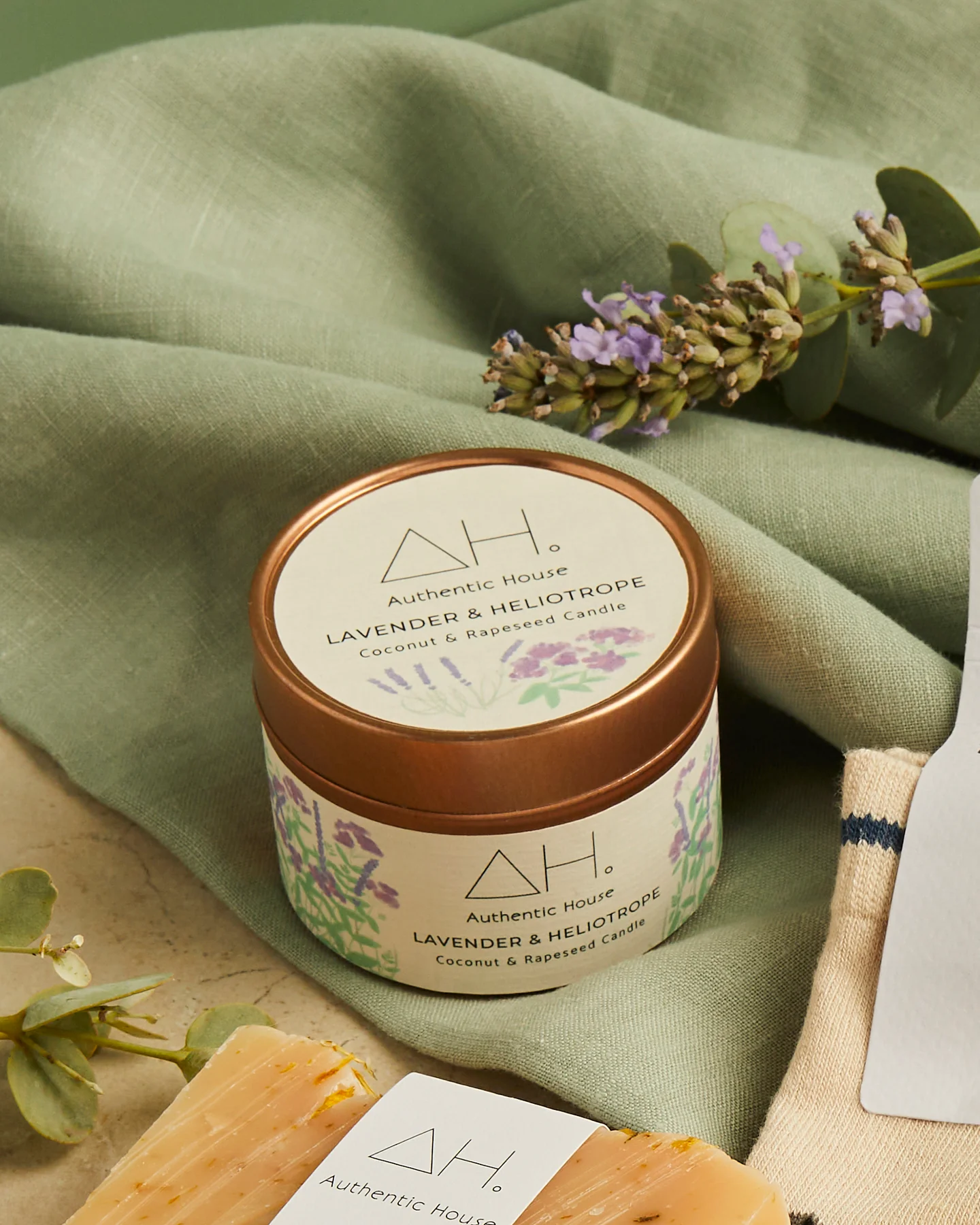 Vegan lavender & heliotrope scented candle in a rose gold coloured tin with lid. Pictured on green fabric with a lavender sprig.