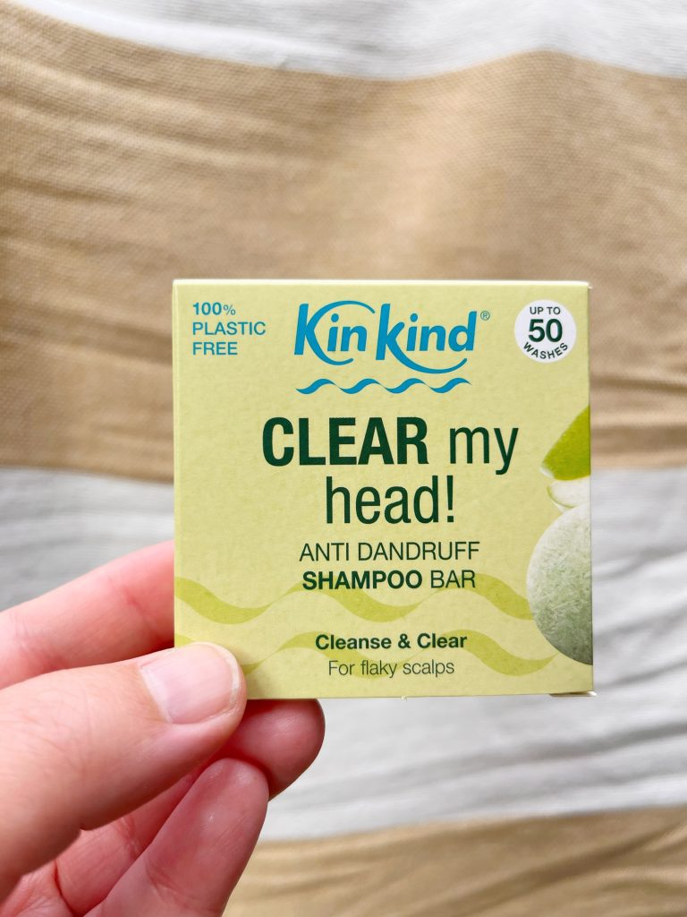 KinKind's 'Clear my Head' anti dandruff shampoo bar for itchy scalps. A hand is holding the small square green box up against a white and beige striped background.