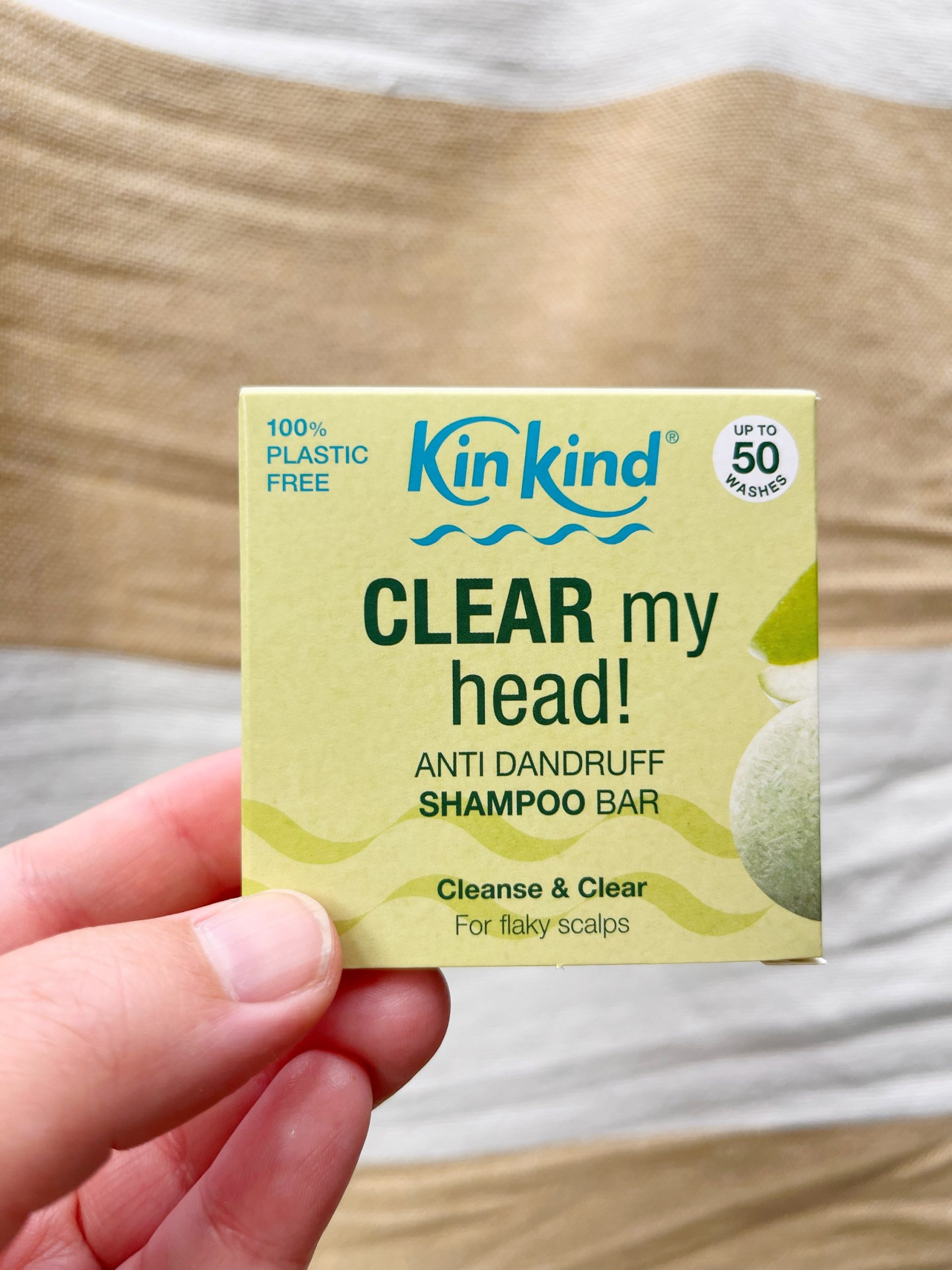 KinKind's 'Clear my Head' anti dandruff shampoo bar for itchy scalps. A hand is holding the small square green box up against a white and beige striped background.