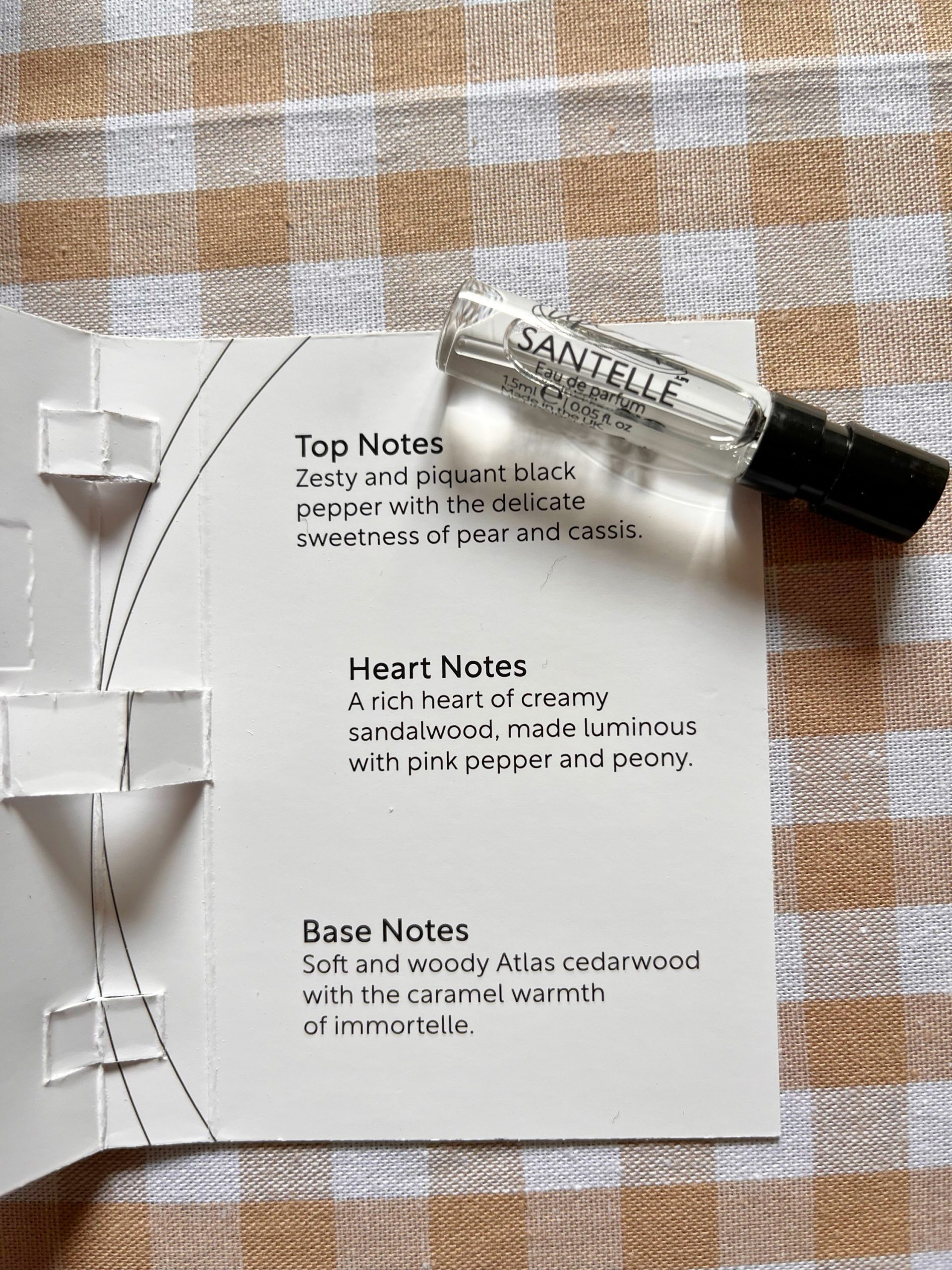 A mini 1.5ml vial of Upcircle's Santelle eau de parfum laying on the white card packaging against a beige gingham background. Available at Sage Folk