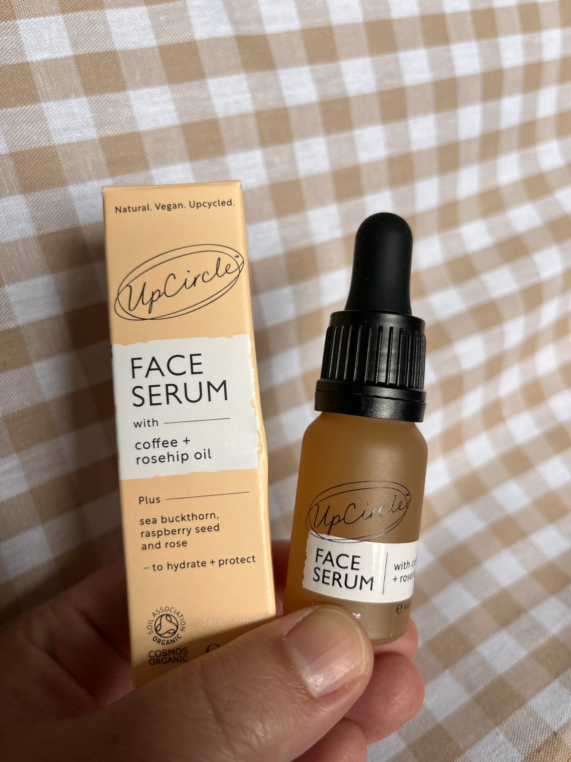 A small orange bottle of Upcircle's organic face serum with a black pippette top. The bottle is being held with the pastel orange cardboard packing. The backdrop is beige gingam material