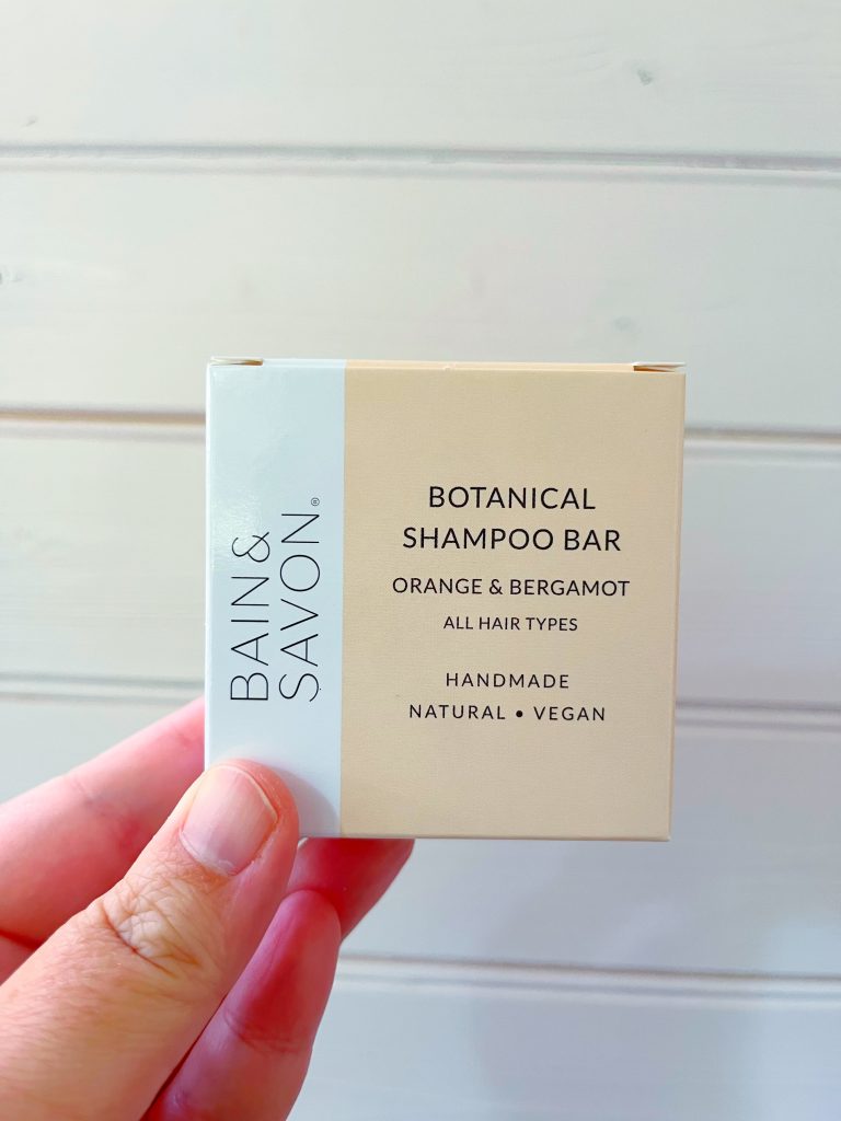 An orange and bergamot shampoo bar designed for all hair types by Bain & Savon. The image shows the square packaging of the shampoo bar (which is pale orange with a white section running along the left edge) being held up against a pale wooden background by a hand.
