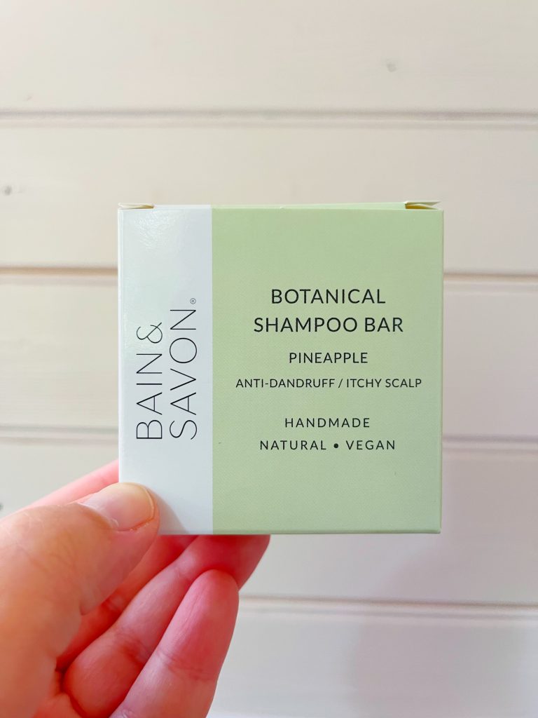 A Pineapple anti dandruff/ itchy scalp shampoo bar by Bain & Savon. The image shows the square packaging of the shampoo bar (which is pale green with a white section running along the left edge) being held up against a pale wooden background by a hand.