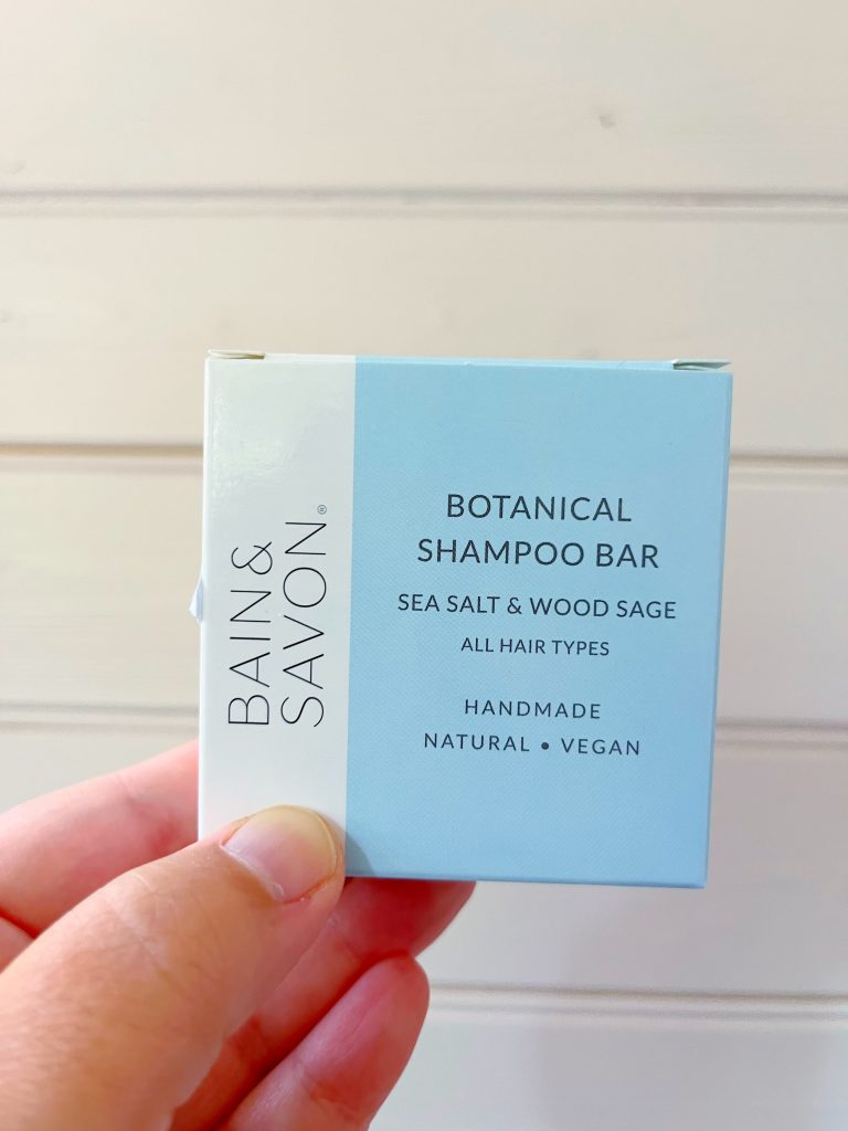 A Sea salt and wood sage shampoo bar designed for all hair types by Bain & Savon. The image shows the square packaging of the shampoo bar (which is light blue with a white section running along the left edge) being held up against a pale pink wooden background by a hand.