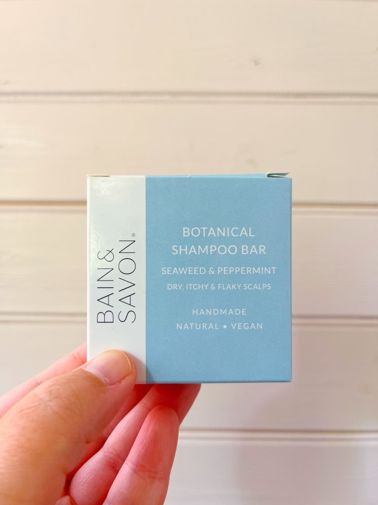 A Seaweed & Peppermint shampoo bar designed for dry, itchy & flaky scalps by Bain & Savon. The image shows the square packaging of the shampoo bar (which is sky blue with a white section running along the left edge) being held up against a pale pink wooden background by a hand.
