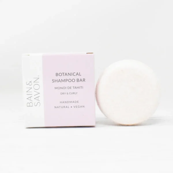 A Monoi De Tahiti shampoo bar designed for dry & curly hair by Bain & Savon. The image shows the square packaging of the shampoo bar (which is pale pink with a white section running along the left edge) with the round shampoo bar next to it. The shampoo bar has a very faint tint of pink to it and the bar and the packaging are against a white background.