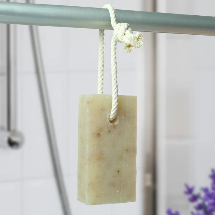 Friendly Soap Lavender natural Soap on a Rope. The bar is a pale off-white colour with darker flecks from the pieces of lavender flower. It's pictured hanging from a metal pole by the white rope cord loop that goes through the top of the bar. There is a small amount of purple lavender flowers visible in the bottom right corner of the photo and a pale tiled background behind the hanging soap bar.