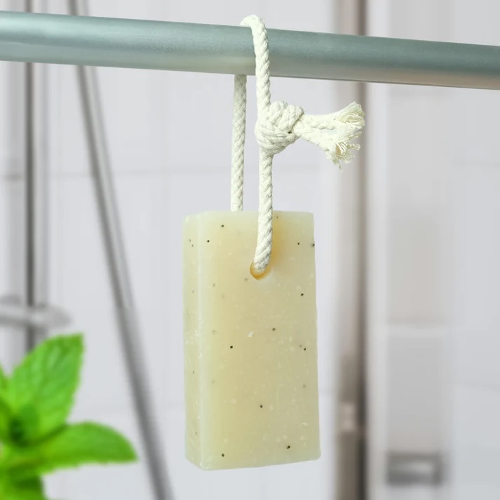 Friendly Soap Peppermint & Poppyseed natural Soap on a Rope. The bar is a pale off-white colour with darker flecks visible from the dried poppy seeds. It's pictured hanging from a metal pole by the white rope cord loop that goes through the top of the bar. There is a small amount of green mint leaves visible in the bottom left corner of the photo and a pale tiled background behind the hanging soap bar.