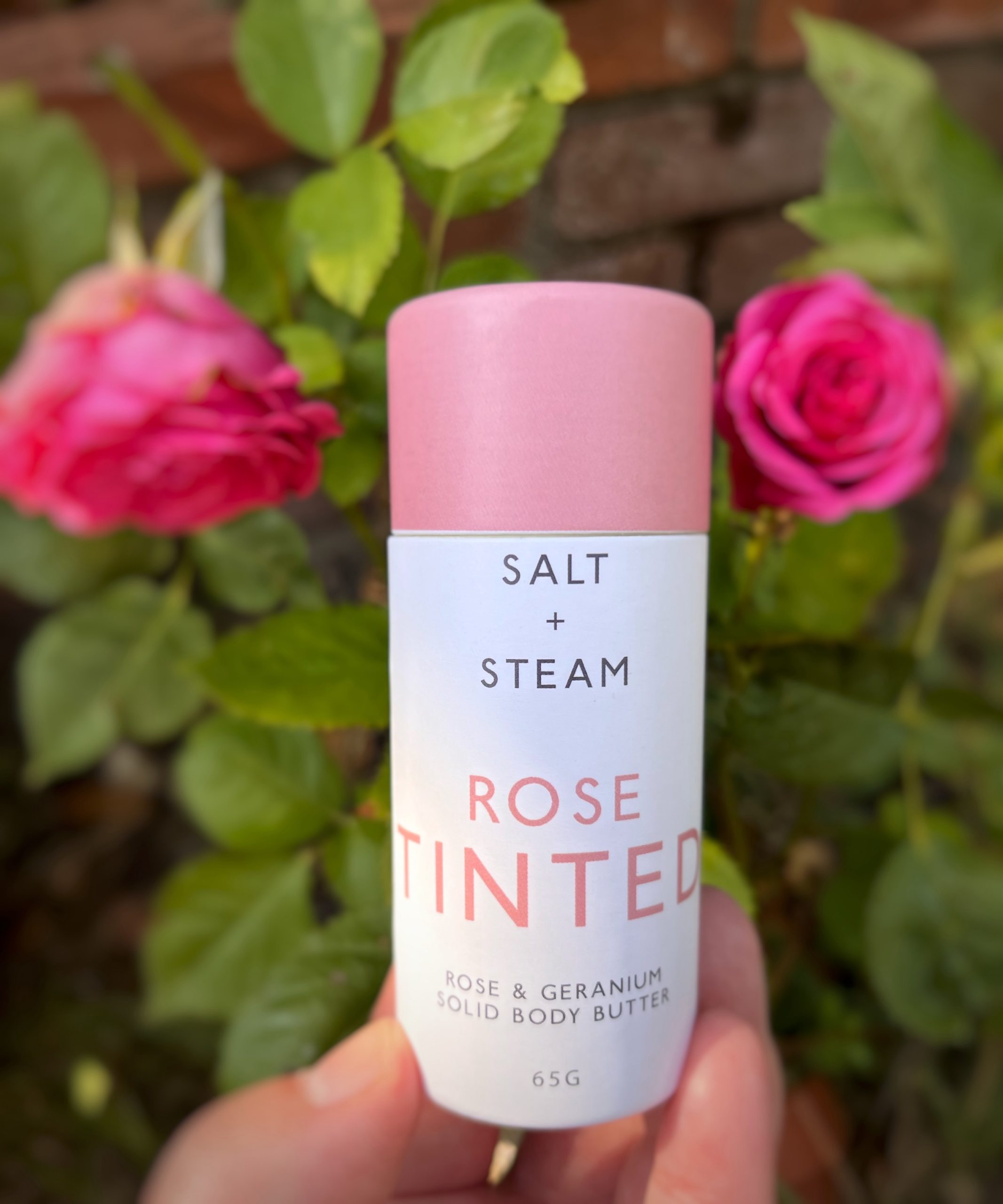 A white cardboard tube with rose pink coloured lid is being held over a gree leafy background with two bright pink roses either side of it